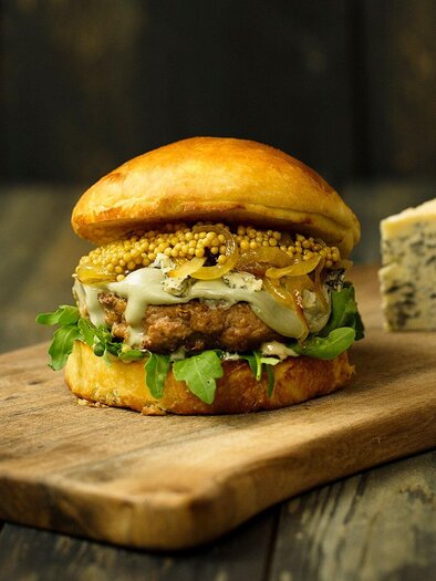 Ein saftiger Burger auf einem Holzbrett, belegt mit Salat, Zwiebeln und Senf. Neben dem Burger liegt ein Stück Käse. | © Freigeist - TG-Graphics