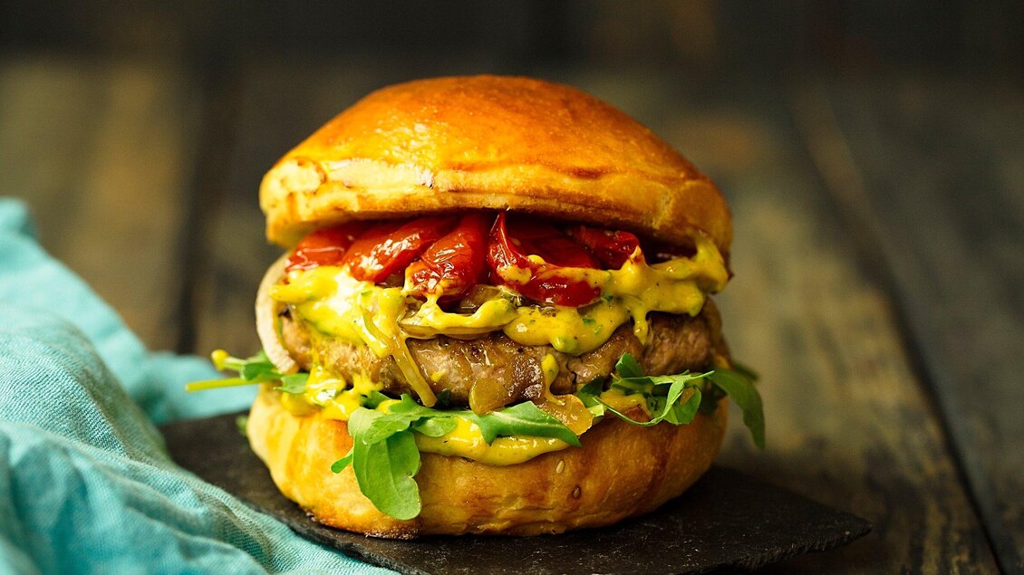 Ein saftiger Burger mit Rindfleisch, schmelzendem Käse, Rucola und gegrillten Tomaten. Serviert auf einem knusprigen Brötchen auf einem dunklen Untergrund. | © Freigeist - TG-Graphics