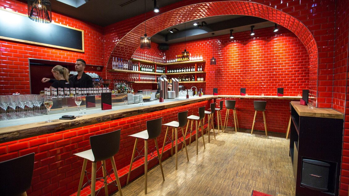 Eine stilvolle Bar mit roten Fliesen und Holzregalen. Im Hintergrund stehen zwei Personen und einige Gläser auf der Theke. | © Freigeist - TG-Graphics