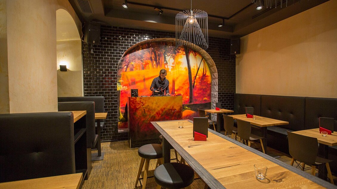 Ein modernes Restaurant mit Holztischen und bequemen Sitzen. An der Wand befindet sich ein auffälliges Wandgemälde und ein DJ-Pult. | © Freigeist - TG-Graphics