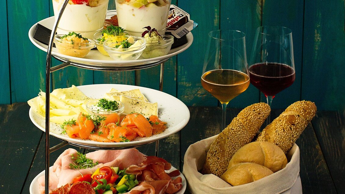 Ein stilvolles Plattenarrangement mit verschiedenen Speisen, darunter Lachs, Aufschnitt und frisches Gemüse. Dazu zwei Gläser Wein und Brötchen in einer Stofftasche. | © Freigeist - TG-Graphics