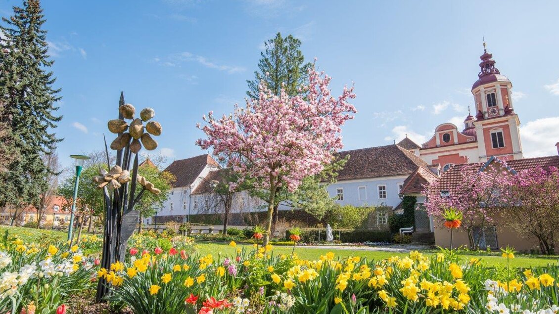 Ein wunderschöner Garten mit bunten Blumen und einem kirschblühenden Baum. Im Hintergrund steht ein charmantes Gebäude mit einem kleinen Turm. | © Helmut Schweighofer