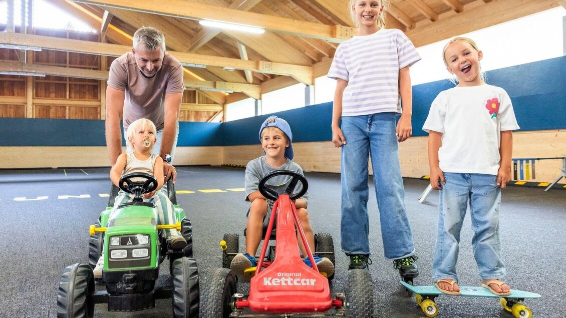 Eine Gruppe von Kindern spielt in einer Halle mit verschiedenen Spielfahrzeugen. Ein Erwachsener steht hinter ihnen und lächelt. | © www.zloam.at