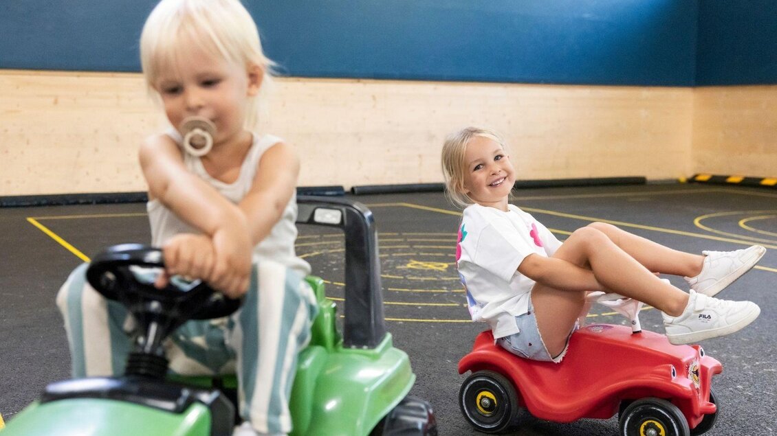 Zwei Kinder fahren mit kleinen Spielzeugautos in einer Innenhalle. Das eine Kind sitzt auf einem grünen Traktor, während das andere Kind auf einem roten Auto lächelt. | © www.zloam.at
