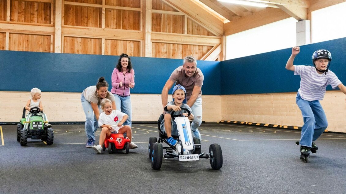 Eine Gruppe von Kindern fährt mit Karts in einer Indoor-Spielumgebung. Ein Erwachsener schiebt ein Kind auf einem Kart, während andere Kinder im Hintergrund spielen. | © www.zloam.at