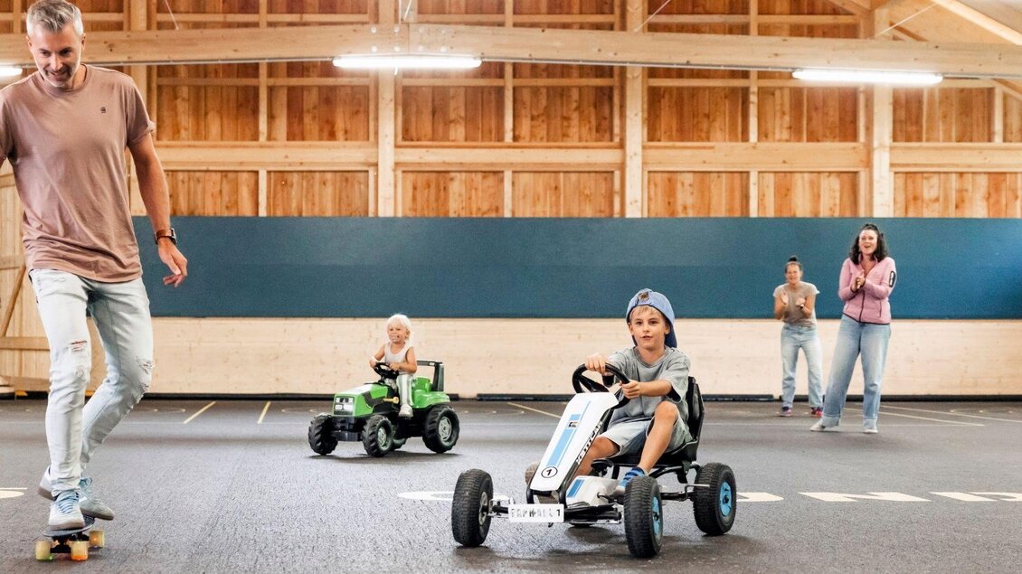 Eine lebhafte Indoor-Szene mit zwei Kindern auf kleinen Fahrzeugen und einem Mann auf einem Skateboard. Im Hintergrund stehen Erwachsene, die zuschauen und lachen. | © www.zloam.at