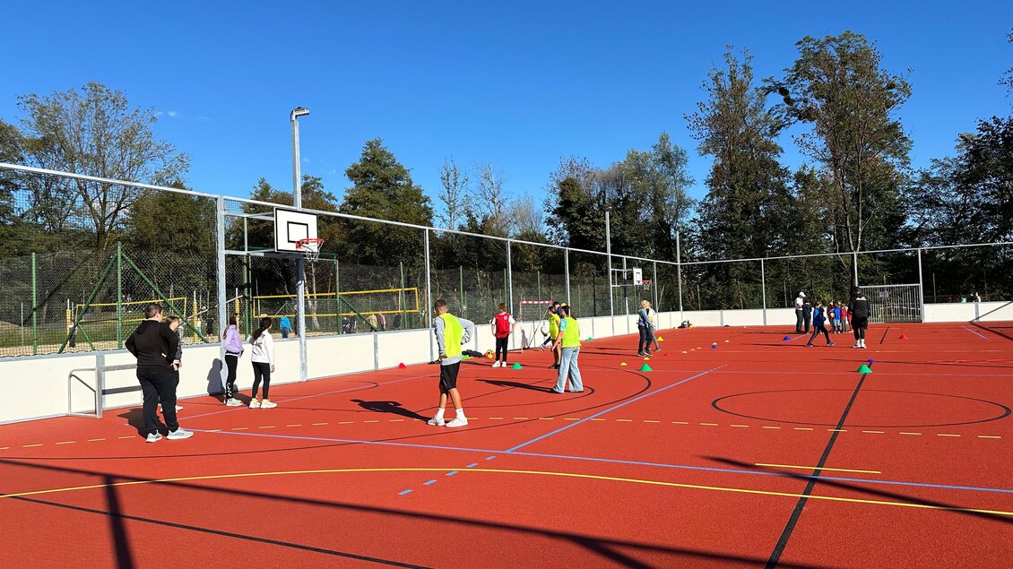 Eine lebhafte Sportanlage mit einem Basketballkorb. Mehrere Personen spielen und trainieren auf dem roten Platz unter klarem Himmel. | © Stadtgemeinde Deutschlandsberg | E. Kleindienst