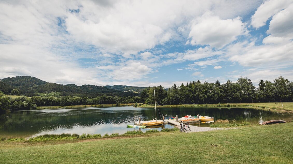 Ein ruhiger See mit Booten und grüner Wiese. Im Hintergrund sind sanfte Hügel und ein schöner Himmel zu sehen. | © nicoleseiser.at
