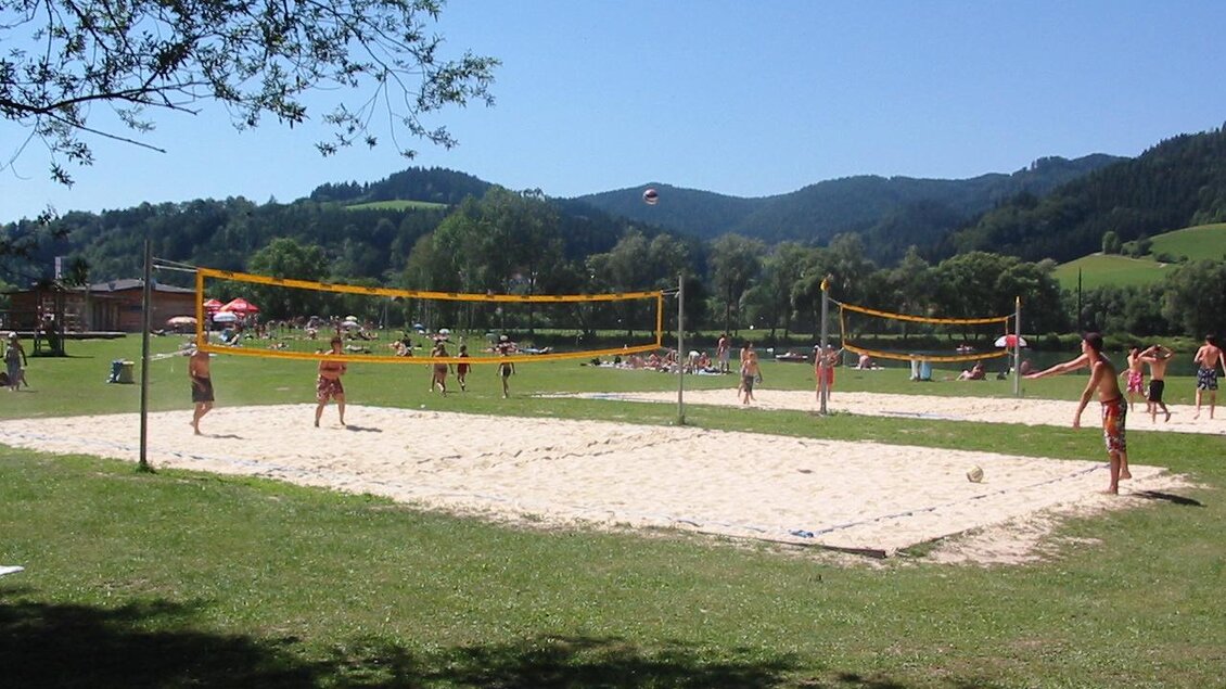 Ein Sandvolleyballplatz in der Natur, umgeben von grünen Hügeln. Mehrere Spieler haben Spaß beim Volleyballspielen unter klarem Himmel. | © Gemeinde Krieglach