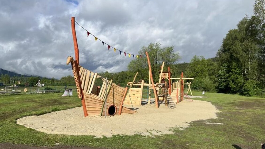 Ein Spielplatz in Form eines Piratenschiffs steht auf einem Sandfeld. Im Hintergrund sind grüne Bäume und ein bewölkter Himmel zu sehen. | © Gemeinde Krieglach