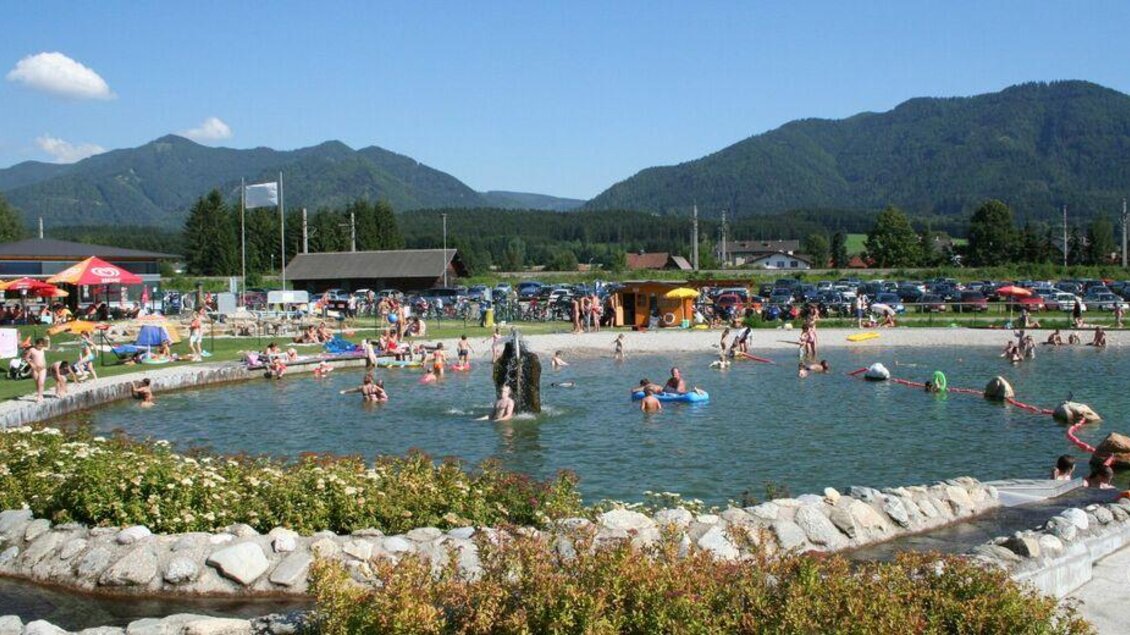 Ein belebtes Schwimmbecken mit vielen Menschen, die relaxen und spielen. Im Hintergrund sind grüne Berge und Sonnenschirme zu sehen. | © Gemeinde Krieglach