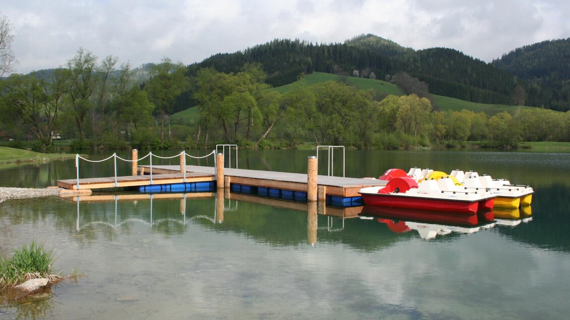 Ein ruhiger See mit einem Holzsteg und einer Bootsanlegestelle. Im Hintergrund sind grüne Hügel und ein bewölkter Himmel zu sehen. | © Gemeinde Krieglach