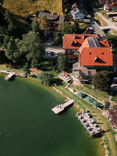 Ein malerisches Gebäude am Ufer eines ruhigen Sees, umgeben von grünen Hügeln. Das Wasser spiegelt die bunten Häuser und die Landschaft wider. | © Christoph Lukas