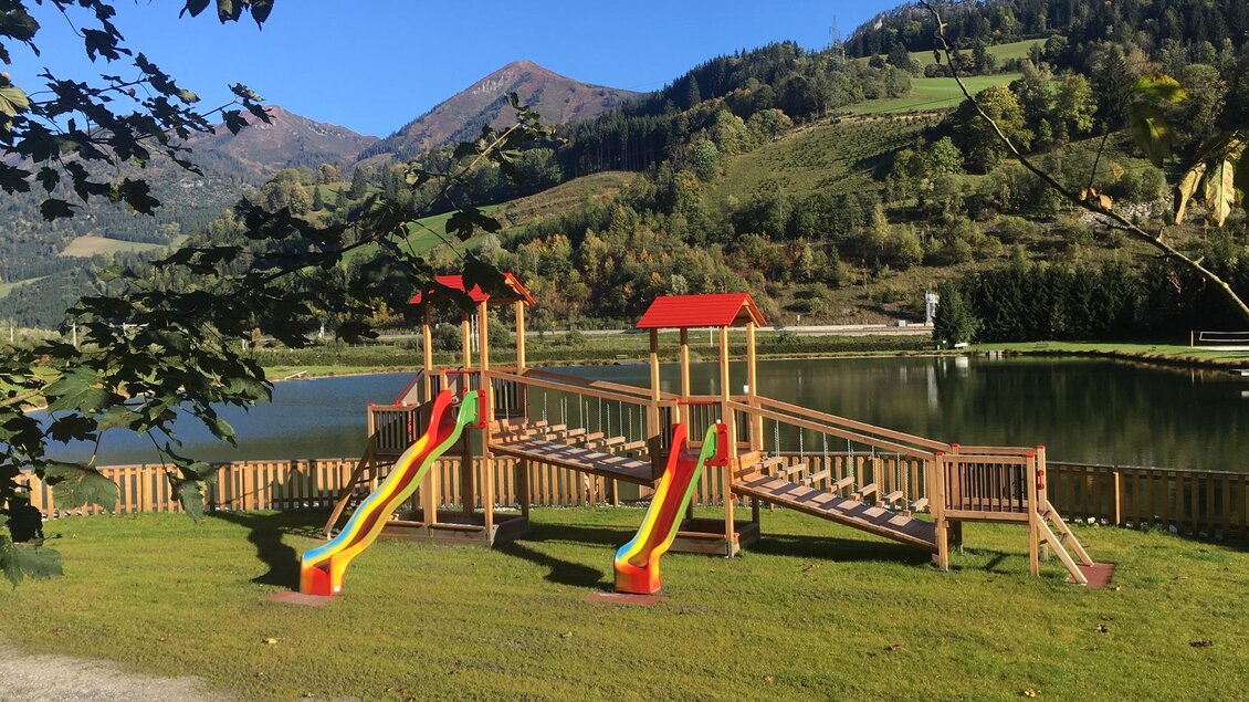 Ein Spielplatz mit Rutschen und einer Holzkonstruktion liegt am Ufer eines klaren Sees. Im Hintergrund sind grüne Hügel und Berge sichtbar. | © Gemeinde Wald am Schoberpass
