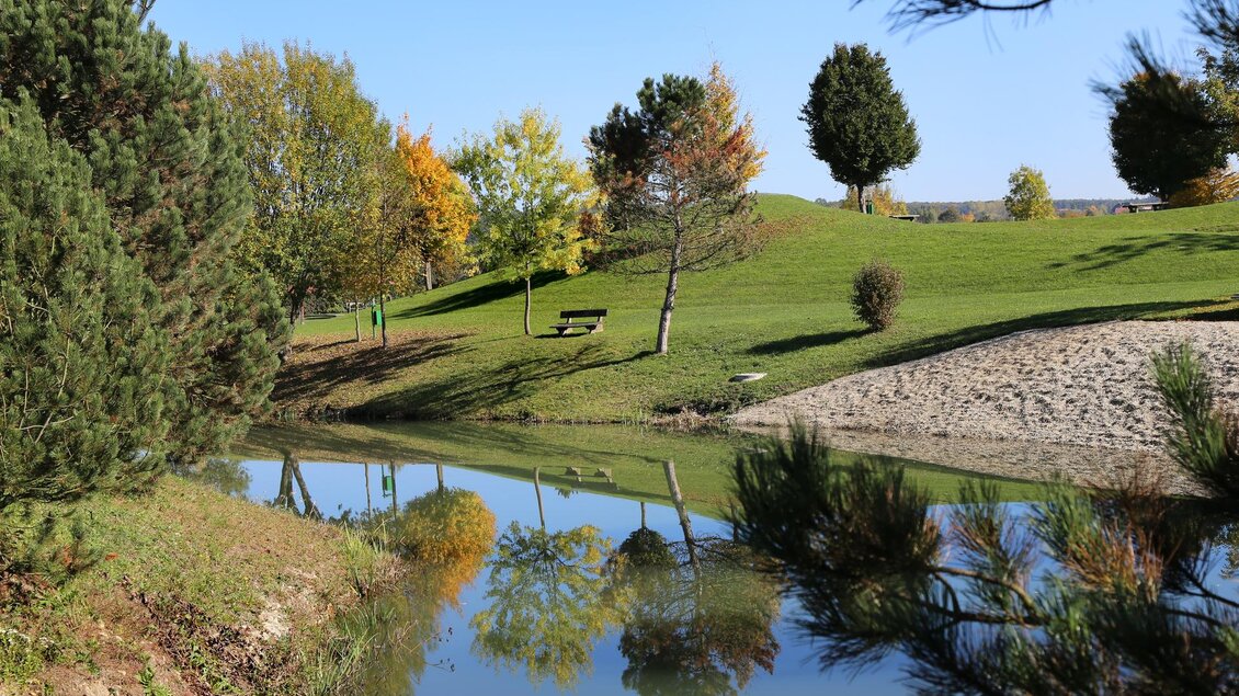 Ein ruhiger Park mit einer spiegelnden Wasserfläche und bunten Bäumen. Im Hintergrund sind Bänke und eine sanfte Hügelkuppen zu sehen. | © Gemeinde Großsteinbach