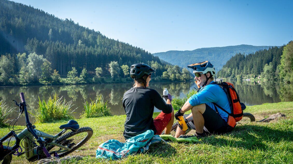 Zwei Radfahrer sitzen am Ufer eines ruhigen Sees und genießen die Landschaft. Im Hintergrund sind Wälder und Berge sichtbar. | © TVB Erzberg-Leoben