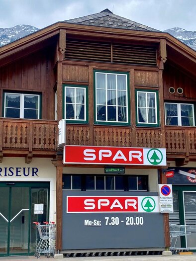 Ein traditionelles Holzgebäude mit einem SPAR-Supermarkt und einem Friseursalon. Im Hintergrund sind Berge zu sehen, und das Wetter ist bewölkt. | © Bettina Scheck