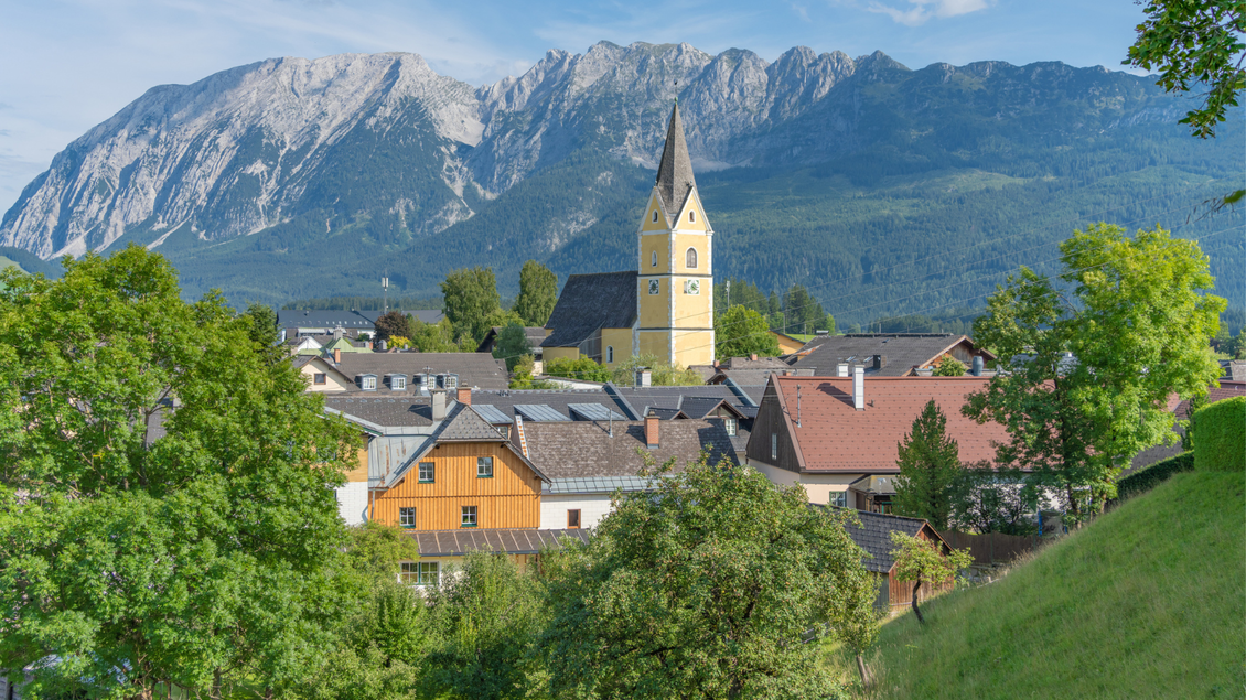 Eine malerische Dorflandschaft mit einer gelben Kirche im Vordergrund. Im Hintergrund erheben sich majestätische Berge unter einem klaren blauen Himmel. | © Manfred Fuchs