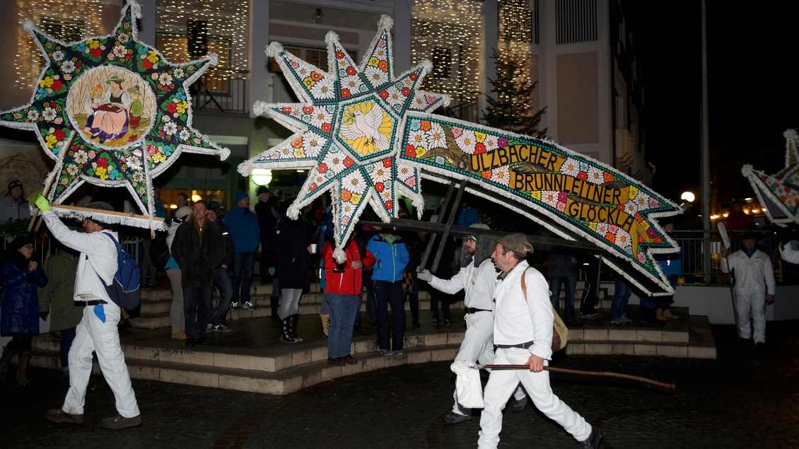 Eine festliche Parade mit Menschen in weißen Anzügen, die große, bunte Sterne tragen. Im Hintergrund stehen Zuschauer und eine beleuchtete Gebäudeansicht. | © Manfred Fuchs