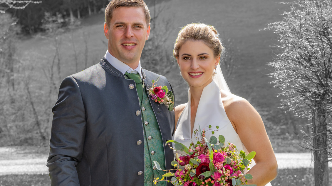 Ein Hochzeitspaar steht im Freien, umgeben von einer schönen Landschaft. Die Braut trägt ein elegantes Kleid und hält einen bunten Blumenstrauß. | © Manfred Fuchs