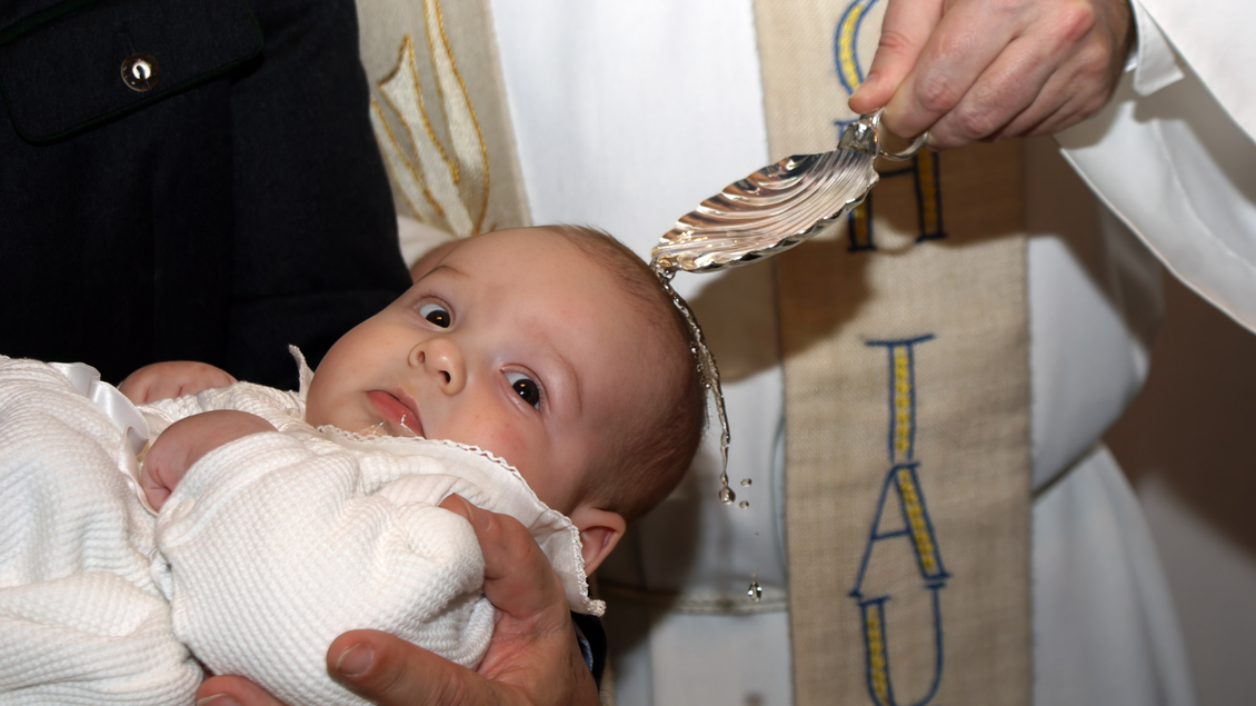 Ein Baby wird während einer Taufe mit Wasser besprengt. Eine Person hält das Baby, während ein Priester das Ritual durchführt. | © Manfred Fuchs