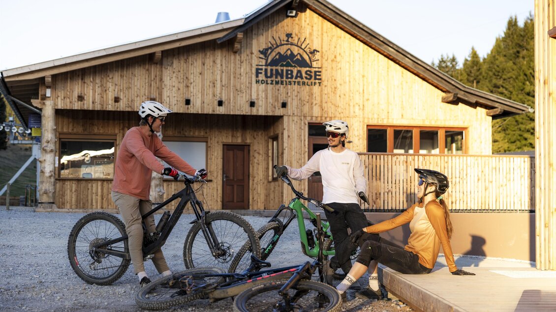 Drei Mountainbiker stehen vor einer Holzhütte mit dem Schriftzug "FUNBASE". Die Szene zeigt eine entspannte Atmosphäre in der Natur. | © Oststeiermark Tourismus