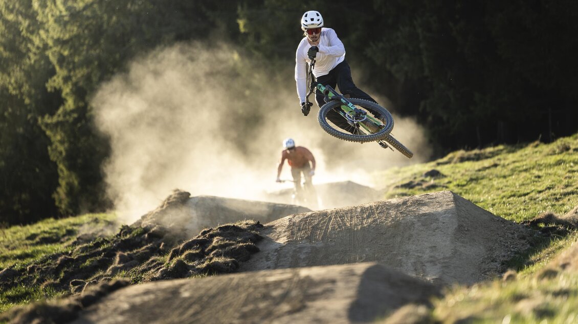 Ein Mountainbiker springt über einen Erdhügel und wirbelt Staub auf. Im Hintergrund ist ein weiterer Fahrer zu sehen. | © Oststeiermark Tourismus