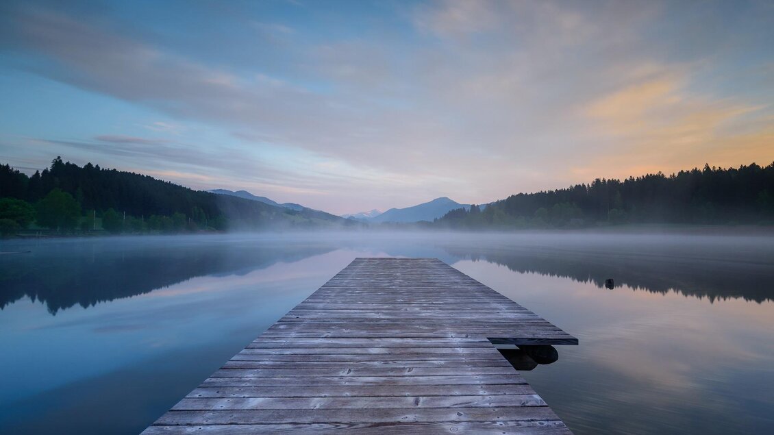 Ein ruhiger See bei Sonnenaufgang mit einem Holzsteg im Vordergrund. Die umliegenden Berge sind leicht im Nebel verborgen. | © Tourismusverband Murau
