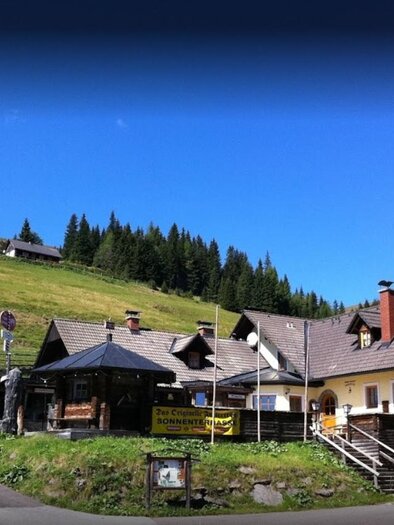 Ein gemütliches Bergrestaurant umgeben von grünen Wiesen und Tannenbäumen. Der Himmel ist blau und klar.