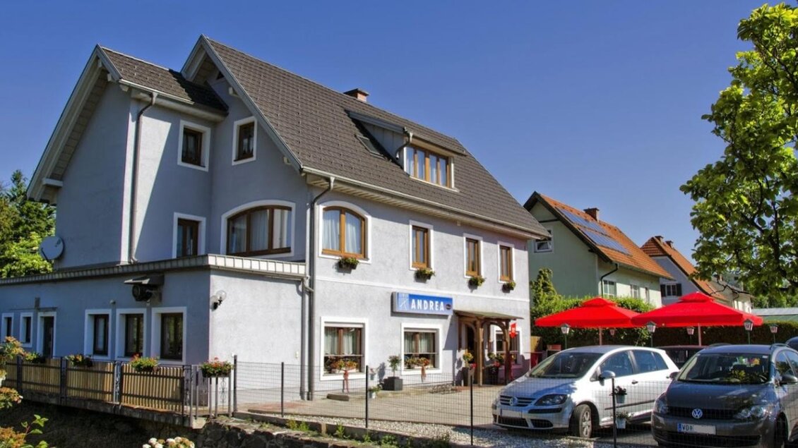 Außenansicht des Gästehauses Café Andrea in Köflach-Pichling mit roten Sonnenschirmen und geparkten Autos vor dem Gebäude. | © Hans Wölkart, Gästehaus Andrea