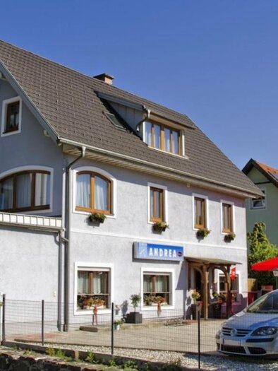 Außenansicht des Gästehauses Café Andrea in Köflach-Pichling mit roten Sonnenschirmen und geparkten Autos vor dem Gebäude. | © Hans Wölkart, Gästehaus Andrea