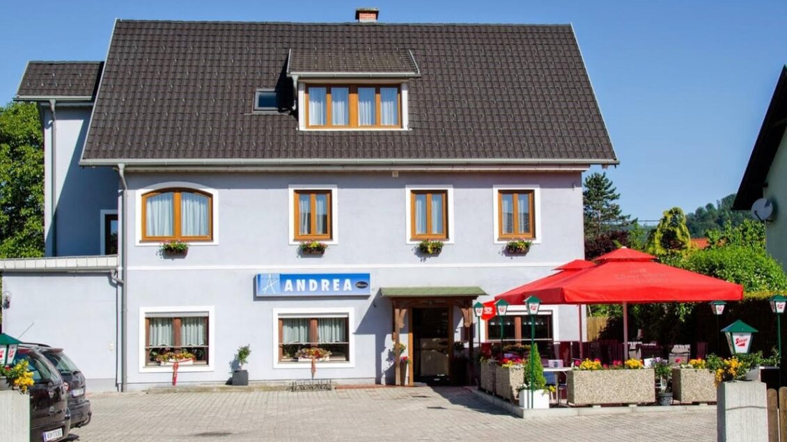 Einladende Außenansicht des Gästehauses Café Andrea in Köflach-Pichling mit roten Sonnenschirmen und Blumenbeeten vor dem Gebäude. | © Hans Wölkart, Gästehaus Andrea
