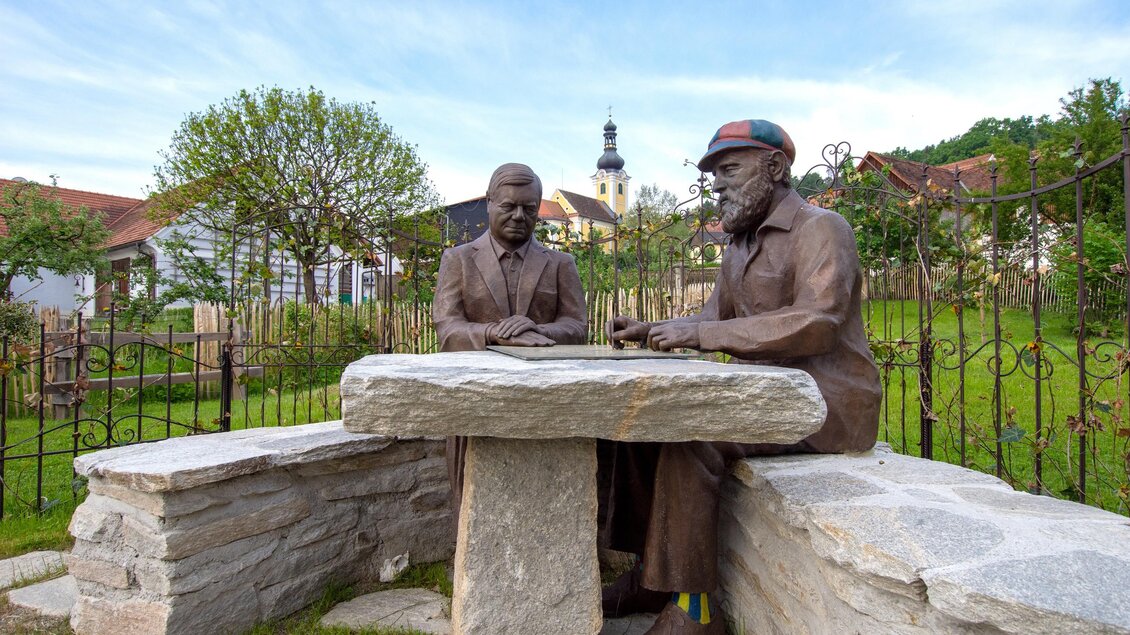 Eine Bronze-Statue von zwei Männern, die an einem Tisch sitzen und ein Spiel spielen. Im Hintergrund sind Bäume und ein Kirchenausblick zu sehen. | © Kurkommission Bad Blumau