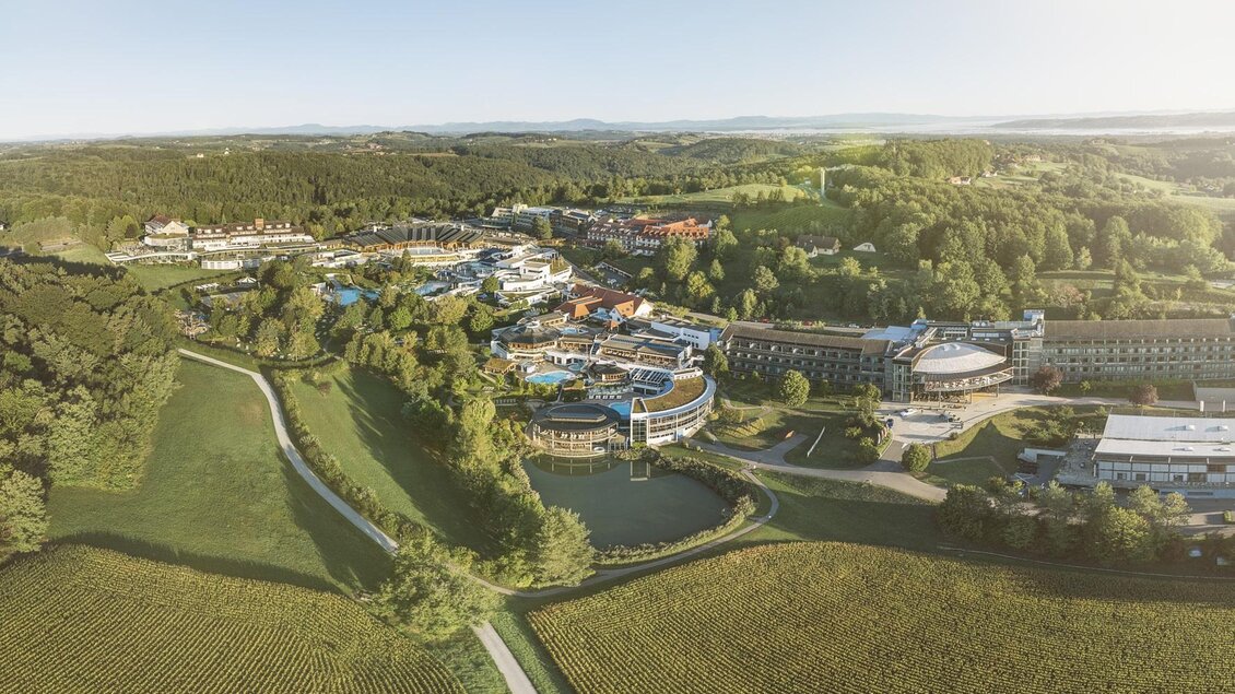 <p>Ein Blick auf eine großzügige Hotelanlage mit Grünflächen und einem kleinen Teich. Die Umgebung ist von sanften Hügeln und Bäumen umgeben. Gästeinfo in Bad Loipersdorf.</p> | © TRL
