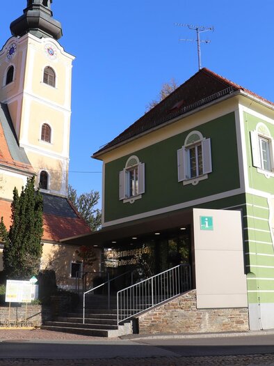 A modern green building stands next to a church with a tall tower. The sky is clear and the surroundings are surrounded by trees. | © Thermen- und Vulkanland
