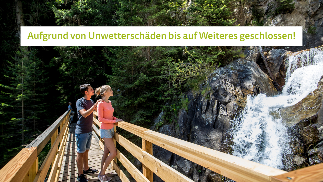 Ein Paar steht auf einer Holzbrücke in einem Wald und betrachtet einen Wasserfall. Die Umgebung ist grün und sonnig mit Bäumen im Hintergrund.