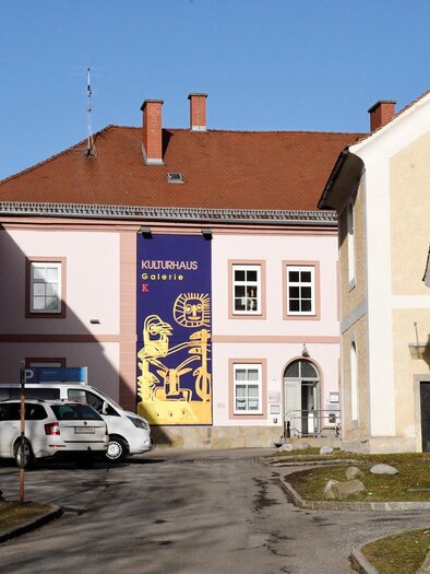 A building with a pink facade and a large banner. A parked car is visible in the foreground, the surroundings are bright and sunny. | © Stadtgemeinde Kindberg