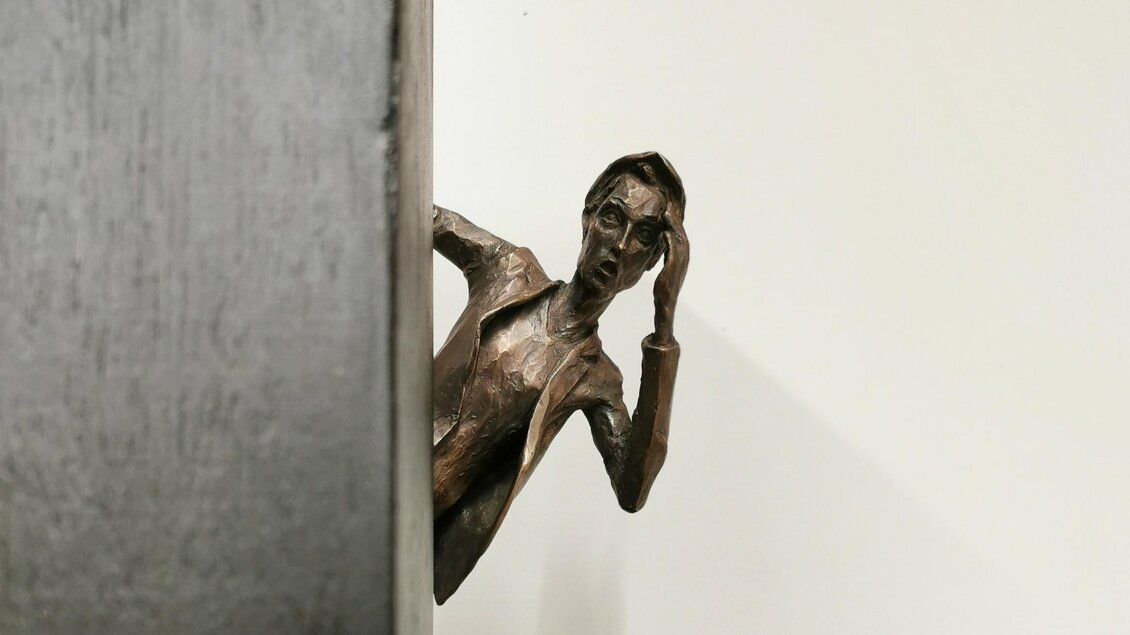 Eine beeindruckende Skulptur eines nachdenklichen Mannes, der hinter einer Wand hervorlugt. Die Figur hat eine bronzene Oberfläche und zeigt eine expressive Haltung. | © Jörg Roskaric
