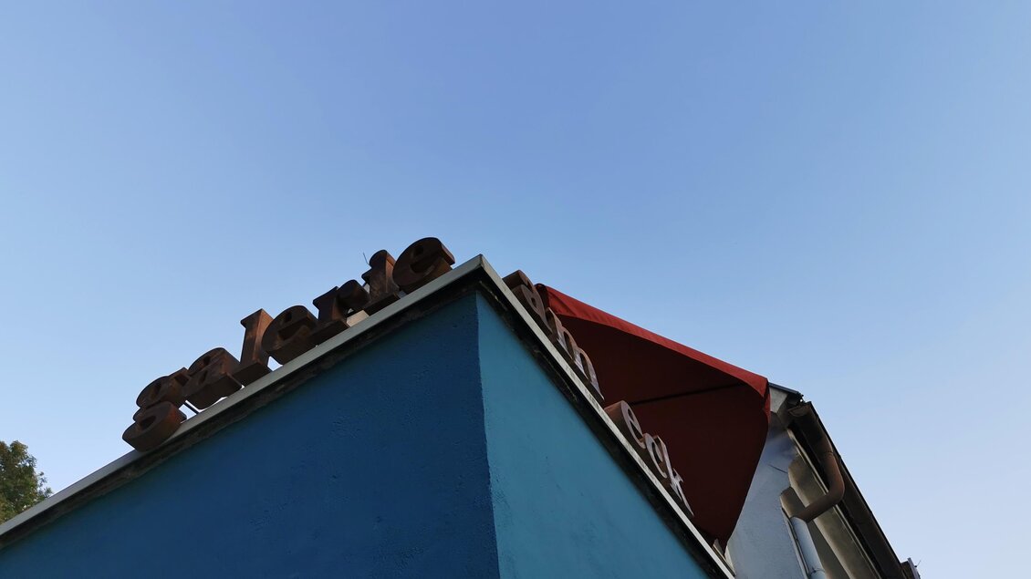 Ein Teil eines Hauses mit einer blauen Wand und einem roten Dachvorsprung. Der Himmel ist klar und blau. | © Jörg Roskaric