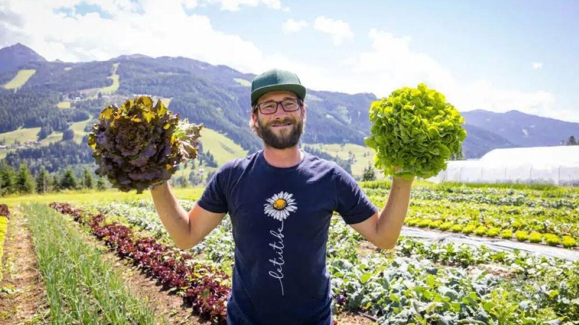 Mann steht im Gemüsefeld und hält zwei große Salatköpfe vor alpiner Landschaft. | © Garten am Berg