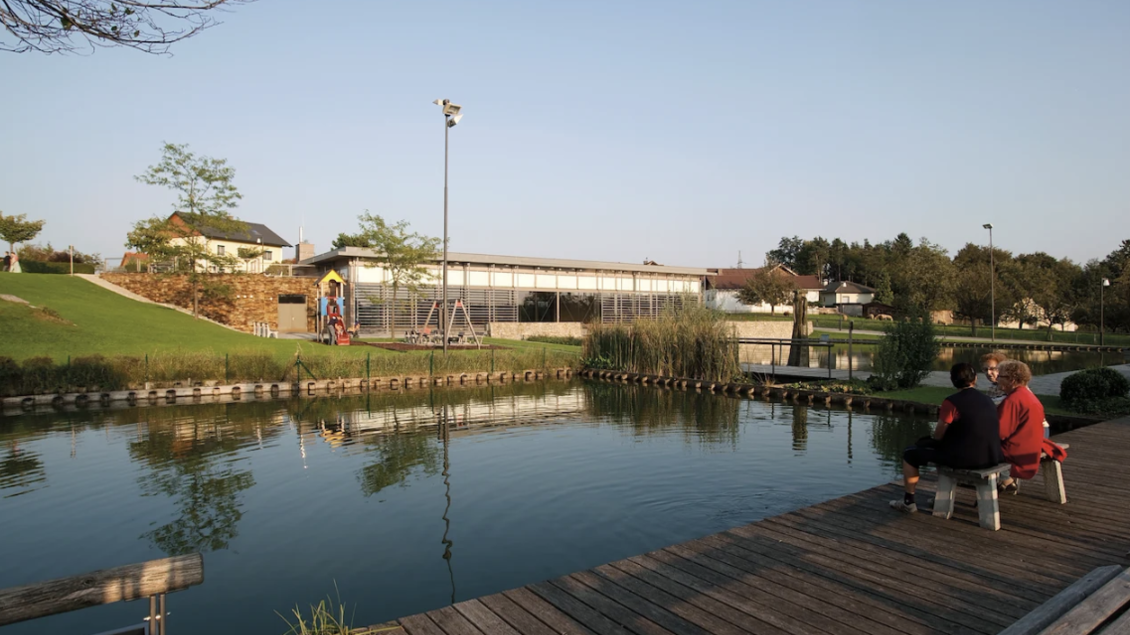 Ein ruhiger Teich mit einer Holzplanke, auf der zwei Personen sitzen. Im Hintergrund sind Bäume und ein Spielplatz zu sehen. | © Oststeiermark Tourismus