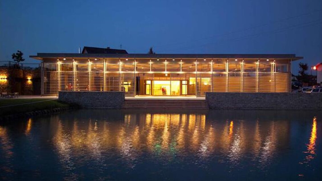 Ein modernes Gebäude am See bei Nacht, beleuchtet von warmem Licht. Die Spiegelung im Wasser verstärkt die beeindruckende Atmosphäre. | © Oststeiermark Tourismus