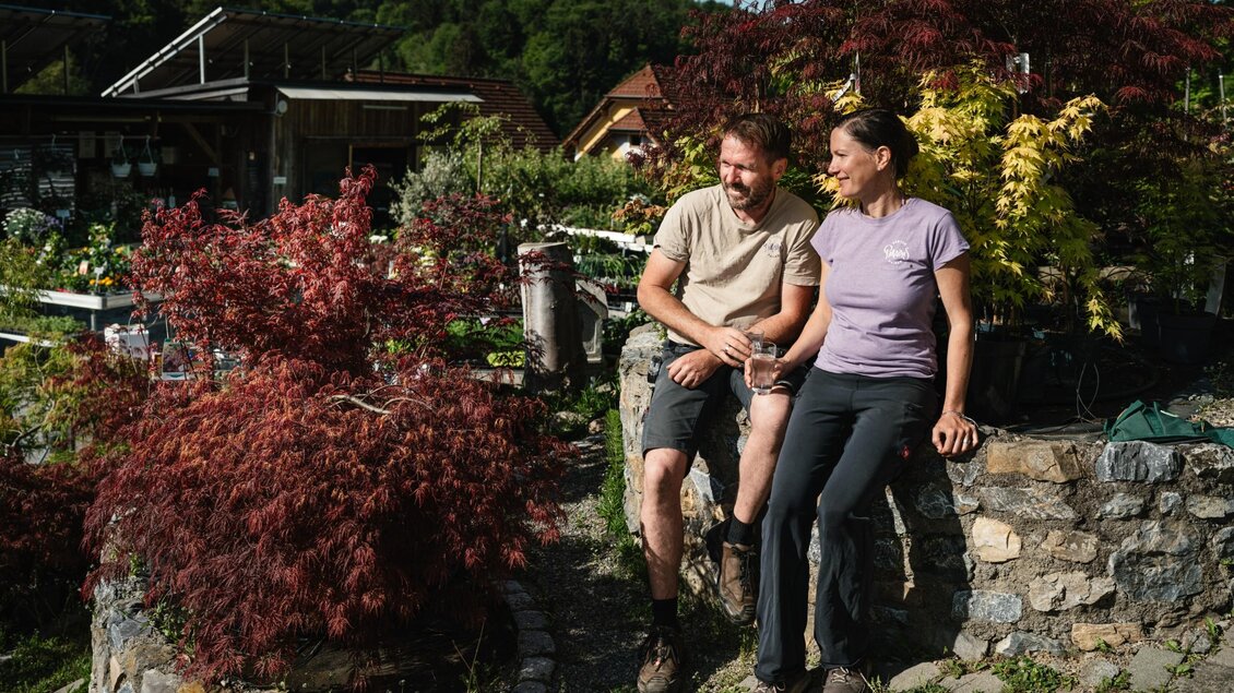 Herr und Frau Painer sitzen auf einer Steinmauer zwischen Ziergehölzen im Freigelände des Gartenparadies Painer. | © Bernd Grosseck