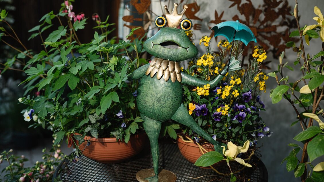 Grüner Deko-Frosch mit Krone und Schirm zwischen blühenden Pflanzen im Gartenparadies Painer. | © Bernd Grosseck