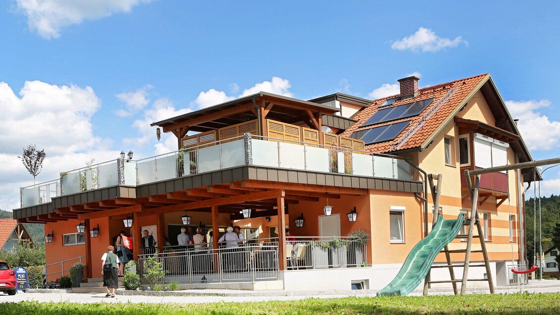 Ein modernes Haus mit einer großen Terrasse und einem grünen Spielplatz. Im Vordergrund sind mehrere Menschen zu sehen, die sich unterhalten. | © TV Region Graz - Lunghammer