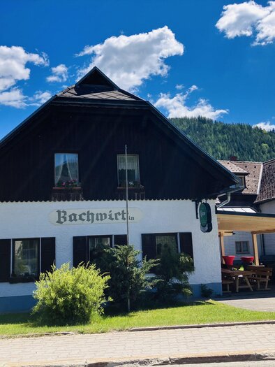 A charming building in rural style with a black roof and an inviting terrace. Surrounded by green plants and a clear blue sky. | © Gasthaus Bachwirt