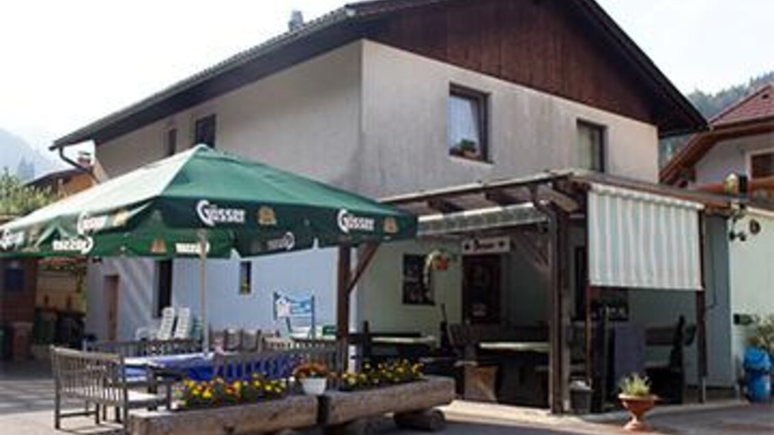 Ein gemütliches Gasthaus mit einem überdachten Außenbereich. Auf der Terrasse stehen Tische mit Blumen und Sonnenschirmen. | © C.Stein