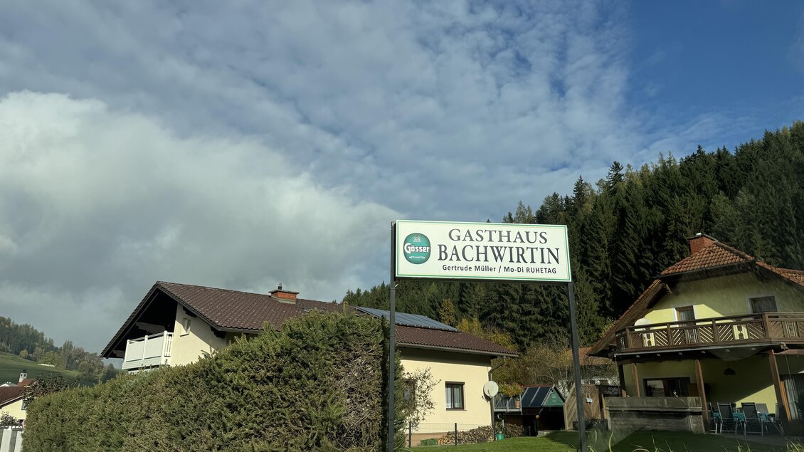 Ein Gasthaus mit dem Namen "Bächwirtin" steht an einer ruhigen Straße. Im Hintergrund sind Bäume und eine bewölkte Himmel sichtbar. | © TV Hochsteiermark