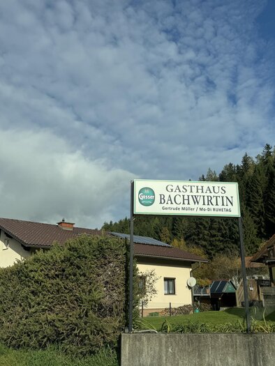 An inn named "Bächwirtin" stands on a quiet street. In the background, trees and a cloudy sky are visible. | © TV Hochsteiermark