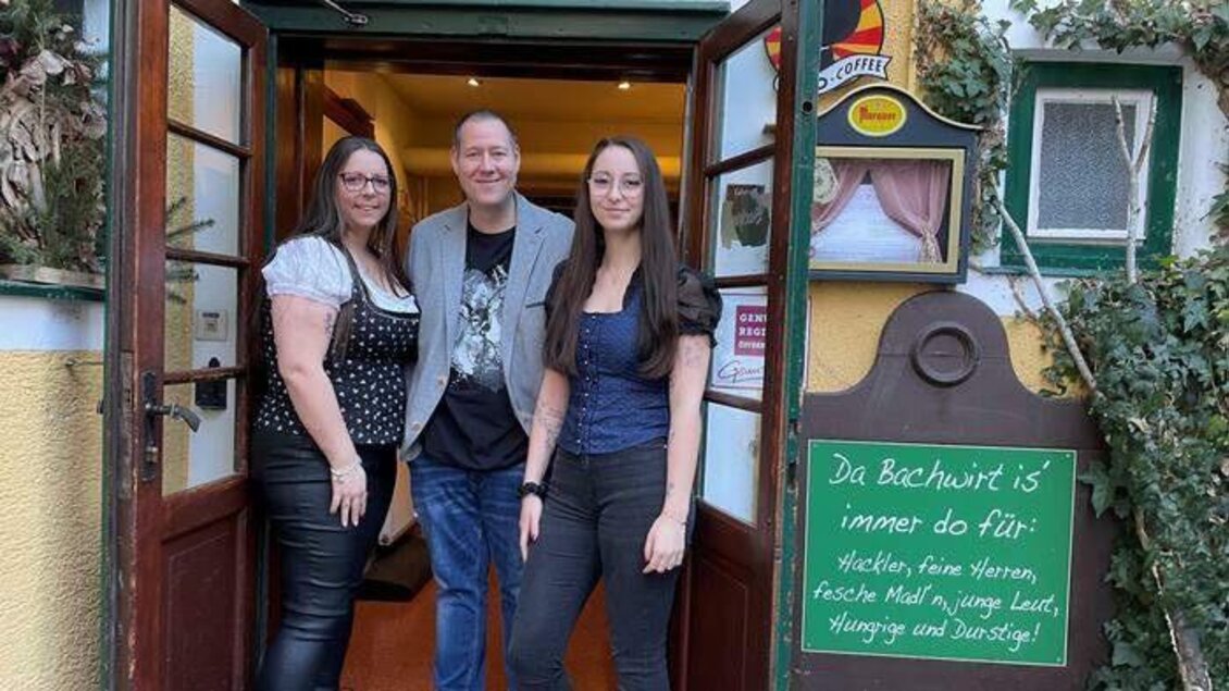 Drei Personen stehen vor einem Restaurant mit einem einladenden Eingang. An der Wand hängt ein Schild mit dem Text „Da Bechwirt is‘ immer do fi‘“. | © Gasthaus Bachwirt